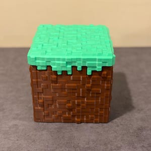 May include: A cube-shaped toy with a pixelated design, featuring a green top representing grass and a brown base resembling earth. The toy is a block, likely from a video game.