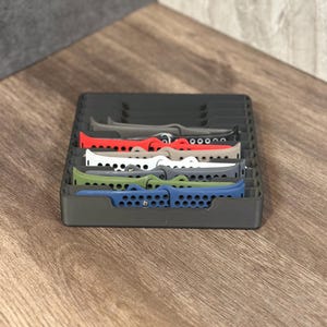May include: A black organizer with six slots for storing watch bands. The bands are in various colors, including red, white, green, blue, and gray.