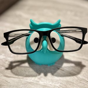 May include: A teal owl-shaped glasses holder with white eyes and black pupils. Black-framed eyeglasses rest on the holder. The holder is made of plastic and sits on a light gray surface.