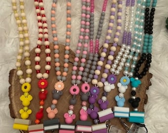 May include: Ten colorful beaded lanyards with different charms, including Mickey Mouse, sunflowers, and flowers. Each lanyard has a clear plastic badge holder attached.