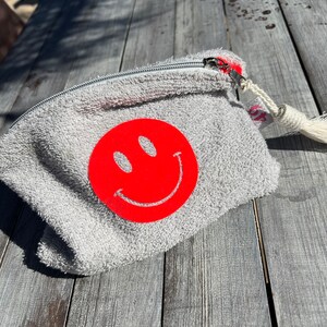 Small cosmetic bag made of light gray terry cloth with a fun neon smiley print and a pretty tassel.