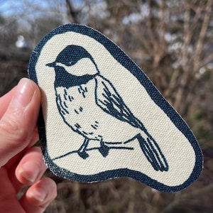 May include: A fabric patch featuring a black-capped chickadee bird illustration. The bird is perched on a branch, with a black head and white body. The patch has a navy blue border and is on a beige background.