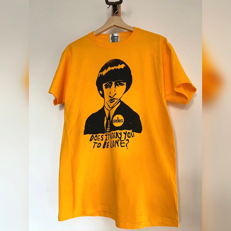 May include: Mustard yellow t-shirt featuring a black graphic of a man in a suit and the text "DOES IT WORRY YOU TO BE ALONE?". The graphic includes the name "B. SHEARS". The shirt is displayed on a wooden hanger.