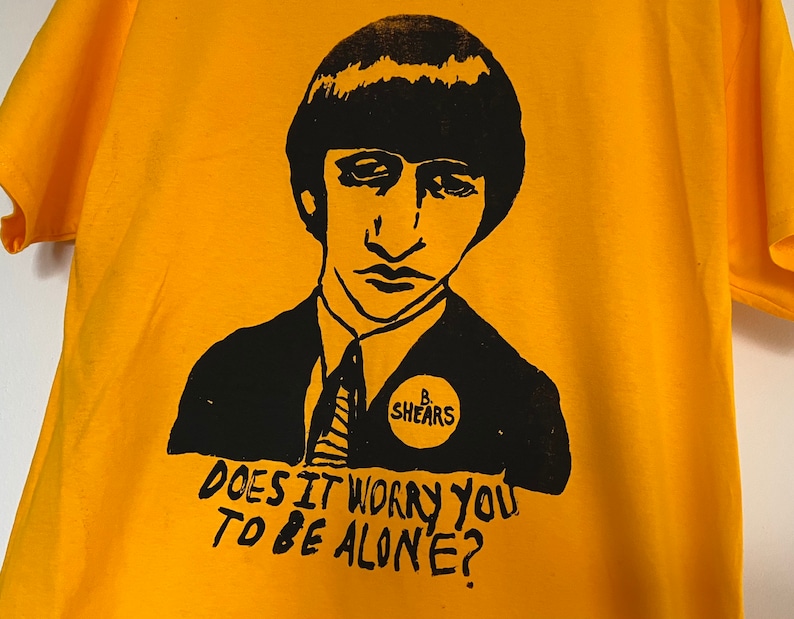 May include: Mustard yellow t-shirt featuring a black graphic of a man's portrait and the text "Does it worry you to be alone?" The man is wearing a suit and tie. A circular badge reads "B. Shears".