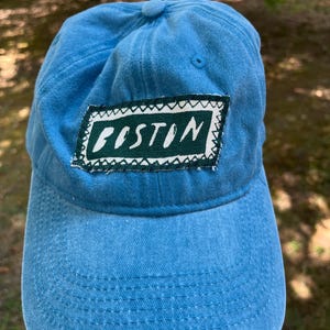 May include: A light blue baseball cap featuring a rectangular dark green patch on the front. The patch has a white border with a zigzag pattern and the word BOSTON printed in white capital letters across the center of the green fabric.
