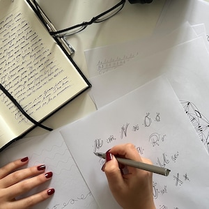 Puede incluir: Una mano escribiendo caligraf&iacute;a en una hoja de papel blanca con un bol&iacute;grafo plateado. El papel tiene un patr&oacute;n de cuadr&iacute;cula y la persona est&aacute; escribiendo letras en cursiva. La persona lleva una chaqueta beige y tiene esmalte de u&ntilde;as rojo en los dedos.