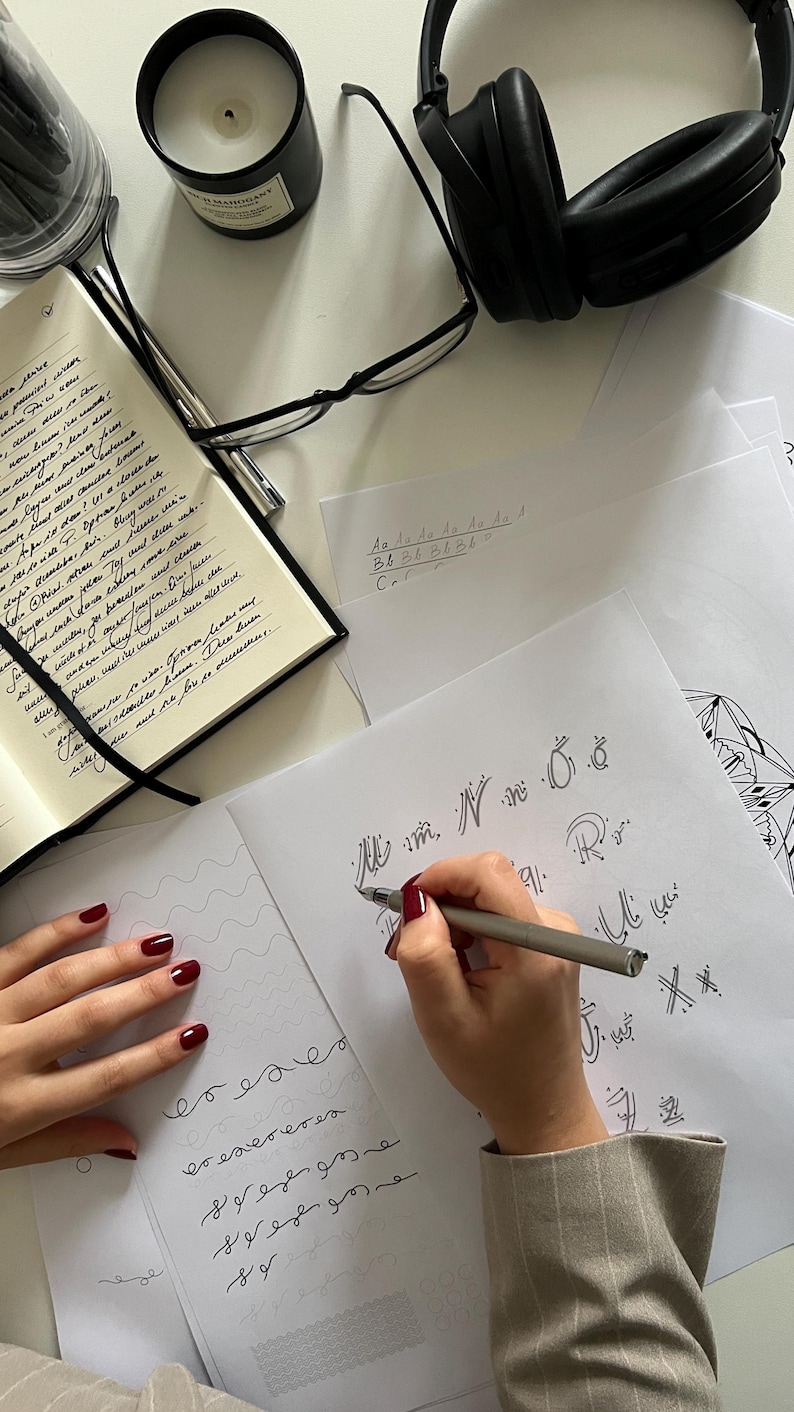 Puede incluir: Una mano escribiendo caligraf&iacute;a en una hoja de papel blanca con un bol&iacute;grafo plateado. El papel tiene un patr&oacute;n de cuadr&iacute;cula y la persona est&aacute; escribiendo letras en cursiva. La persona lleva una chaqueta beige y tiene esmalte de u&ntilde;as rojo en los dedos.