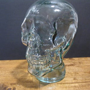 May include: A clear glass skull-shaped decorative object. The skull has detailed facial features, including eye sockets, a nose cavity, and teeth. The glass has a slight green tint and sits on a wooden surface.