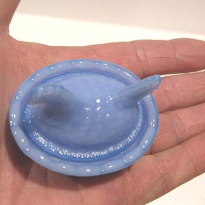 May include: A light blue condom with a reservoir tip, held in a hand. The condom is a pale, translucent blue color and appears to be made of latex. The reservoir tip is a slightly darker shade of blue.