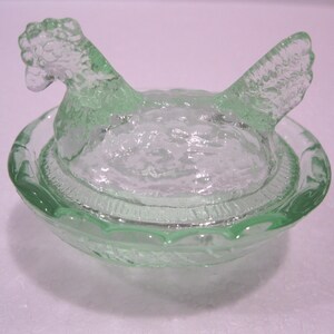 May include: A light green glass hen-shaped dish with a matching lid. The lid is shaped like a hen with its head and wings visible. The dish has a textured surface and is likely used for serving or storage.
