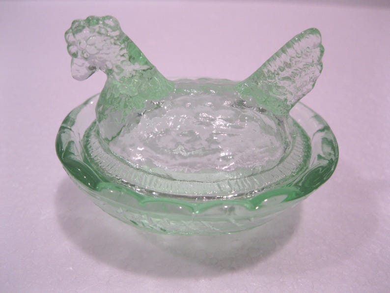 May include: A light green glass hen-shaped dish with a matching lid. The lid is shaped like a hen with its head and wings visible. The dish has a textured surface and is likely used for serving or storage.