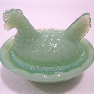 May include: A light green glass hen figurine in a nest-like bowl. The hen has a textured body and a detailed comb and tail feathers. The bowl has a textured rim and a smooth interior. The figurine is a decorative item.
