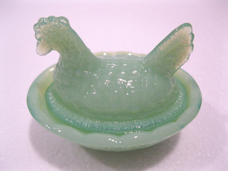 May include: A light green glass hen figurine in a nest-like bowl. The hen has a textured body and a detailed comb and tail feathers. The bowl has a textured rim and a smooth interior. The figurine is a decorative item.