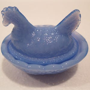May include: A light blue glass hen-on-nest dish. The hen has a textured surface and is perched atop a matching oval-shaped nest. The glass has a glossy finish, reflecting light. This decorative piece is a charming addition to any collection.