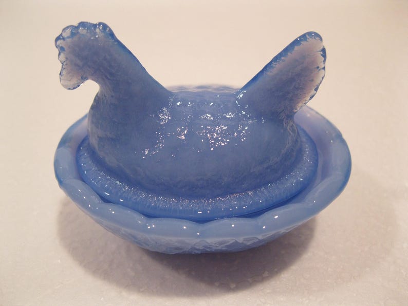 May include: A light blue glass hen-on-nest dish. The hen has a textured surface and is perched atop a matching oval-shaped nest. The glass has a glossy finish, reflecting light. This decorative piece is a charming addition to any collection.