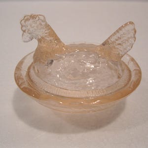May include: A clear glass hen-shaped dish with a lid. The hen is perched atop a round bowl, with a textured surface. The dish is a light peach color and appears to be made of pressed glass.