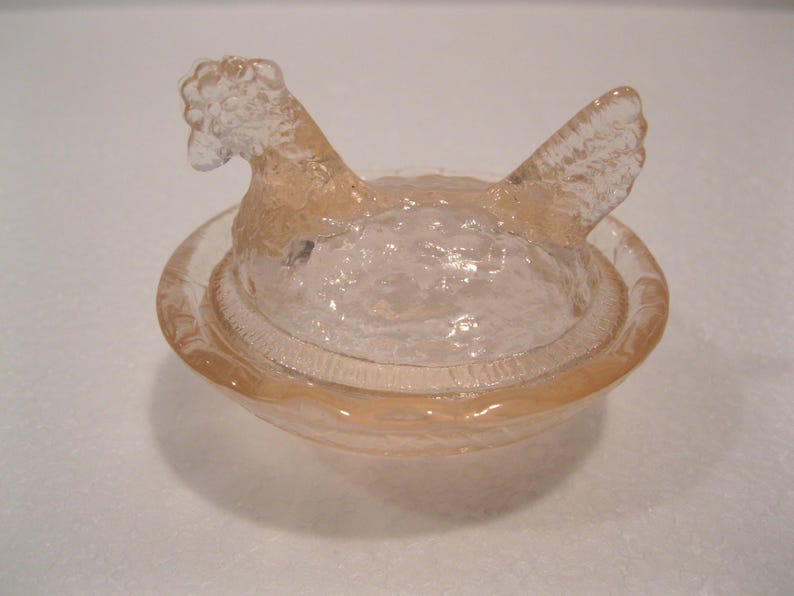 May include: A clear glass hen-shaped dish with a lid. The hen is perched atop a round bowl, with a textured surface. The dish is a light peach color and appears to be made of pressed glass.
