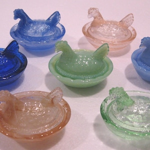 May include: Seven colorful glass hen-shaped trinket dishes. The dishes are in shades of blue, green, and peach. Each hen sits atop a small, round dish. The glass has a textured, translucent appearance.