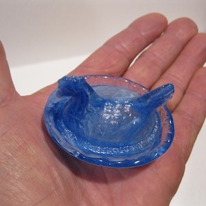 May include: A translucent blue glass object with a circular base and two raised, wing-like structures. The object is held in a hand, showcasing its intricate details and color.