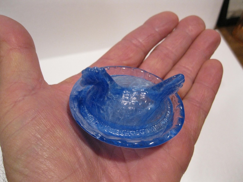 May include: A translucent blue glass object with a circular base and two raised, wing-like structures. The object is held in a hand, showcasing its intricate details and color.