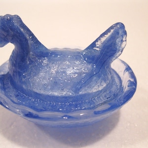 May include: A blue glass hen figurine with a nest-like base. The hen has a detailed head and tail feathers. The glass has a marbled effect with white and blue swirls, creating a unique decorative piece.