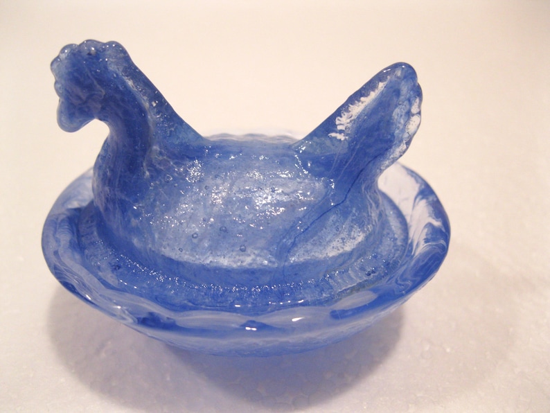 May include: A blue glass hen figurine with a nest-like base. The hen has a detailed head and tail feathers. The glass has a marbled effect with white and blue swirls, creating a unique decorative piece.