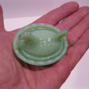 May include: A small, light green glass dish with a textured surface and two raised, curved handles. The dish has a rounded shape with a slightly darker green rim and is held in a person's hand.
