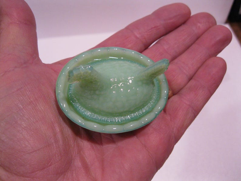 May include: A small, light green glass dish with a textured surface and two raised, curved handles. The dish has a rounded shape with a slightly darker green rim and is held in a person's hand.