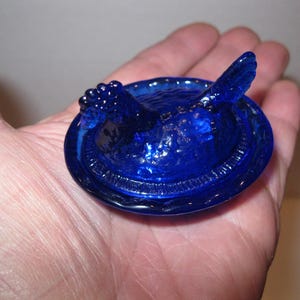 May include: A cobalt blue glass dish with a bird-shaped lid. The lid features a detailed bird design with textured wings and a rounded body. The dish has a circular shape with a decorative rim. The glass has a glossy finish.