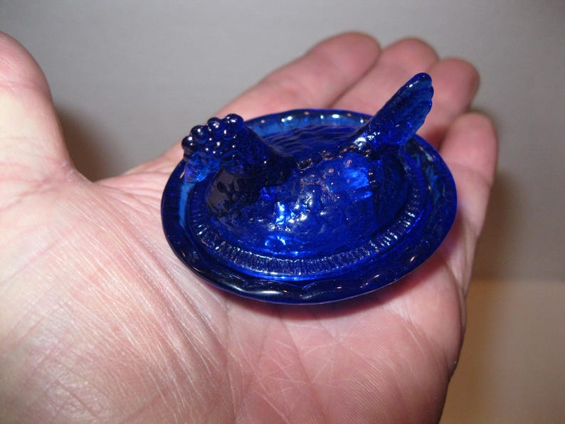 May include: A cobalt blue glass dish with a bird-shaped lid. The lid features a detailed bird design with textured wings and a rounded body. The dish has a circular shape with a decorative rim. The glass has a glossy finish.