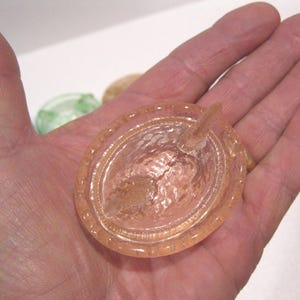May include: A peach-colored glass dish with a textured interior and a small, pointed handle. The dish is oval-shaped with a decorative rim. The image shows the dish held in a hand.
