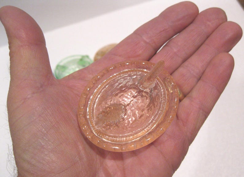 May include: A peach-colored glass dish with a textured interior and a small, pointed handle. The dish is oval-shaped with a decorative rim. The image shows the dish held in a hand.