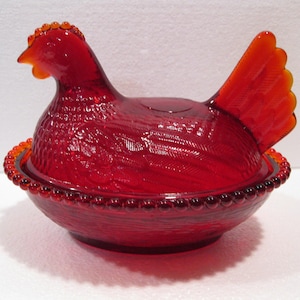 May include: A translucent ruby red glass hen on a nest dish. The lid is shaped like a sitting hen with detailed feather textures, while the base features a beaded rim and a woven basket pattern. The glass is vibrant and catches the light.