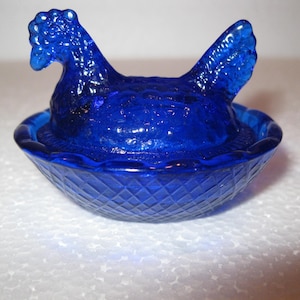 May include: A cobalt blue glass hen on a nest-shaped dish. The hen has a detailed comb and tail feathers. The dish has a diamond pattern on the exterior. A decorative piece.