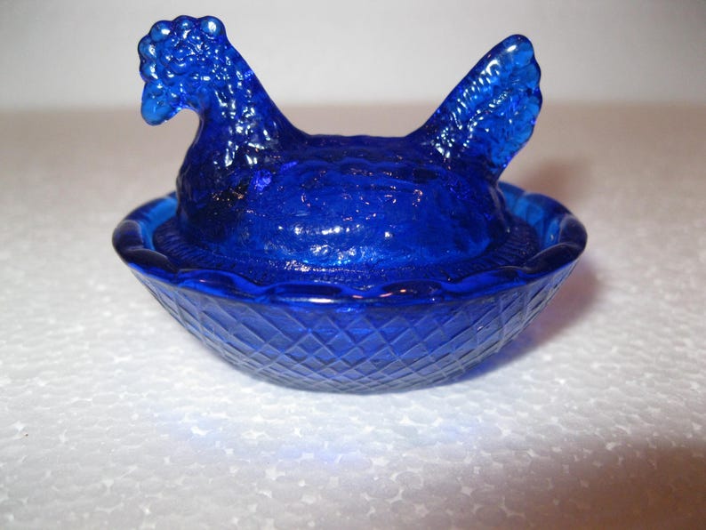 May include: A cobalt blue glass hen on a nest-shaped dish. The hen has a detailed comb and tail feathers. The dish has a diamond pattern on the exterior. A decorative piece.