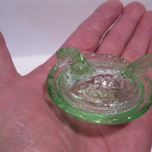 May include: A small, green glass hen-shaped dish with a lid. The dish is round with a detailed rim and the hen has a textured, openwork design. The glass has a slight green tint.