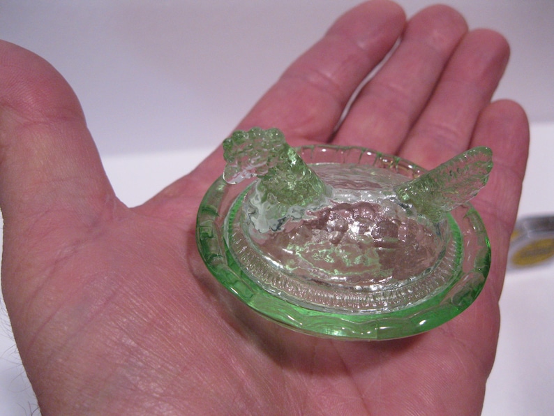 May include: A small, green glass hen-shaped dish with a lid. The dish is round with a detailed rim and the hen has a textured, openwork design. The glass has a slight green tint.
