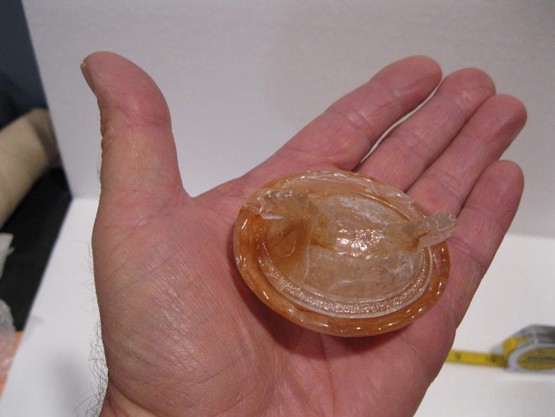 May include: A translucent, amber-colored, oval-shaped object with a raised rim and a small, protruding handle. The object is held in an open hand, showcasing its intricate details and design. The object appears to be made of glass or a similar material.