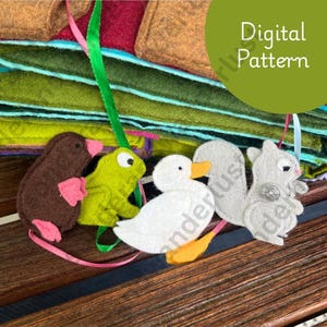 May include: A collection of felt animal ornaments, including a brown mole with pink accents, a green frog, a white duck with an orange beak and feet, and a gray squirrel. The ornaments are attached to colorful ribbons. The image also includes the text "Digital Pattern".