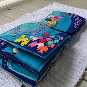 May include: A handmade felt book with an ocean theme. The book features various shades of blue and teal, with colorful felt fish and other sea creatures appliqued on the pages. The book is open, revealing several layers of felt pages.