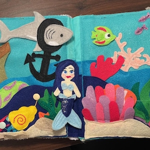 Op de afbeelding: Een vilten stille boek met een oceaanthema. De pagina's tonen zeemeerminnen, een haai, een anker, een zeepaardje, vissen en koraal in verschillende kleuren. Het boek is ontworpen voor kinderen.