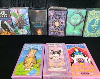 May include: A collection of ten tarot decks with various themes, including crows, cats, spirits, and shadows. The decks are all in different colors and styles, and some feature illustrations of animals.