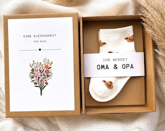 Puede incluir: Una caja de regalo de cartón marrón que contiene un calcetín de bebé blanco con un patrón de osos marrones, envuelto en una banda de papel blanca con el texto IHR WERDET OMA & OPA. Al lado, una tarjeta dice EINE KLEINIGKEIT FÜR EUCH con flores.