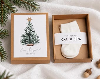 Könnte beinhalten: Eine braune Pappschachtel mit einem weißen Babysöckchen, das von einer Papierbanderole mit dem Text IHR WERDET OMA & OPA umwickelt ist. Der dazugehörige Deckel zeigt ein Aquarell eines Weihnachtsbaums und den Schriftzug Eine Kleinigkeit FÜR EUCH.