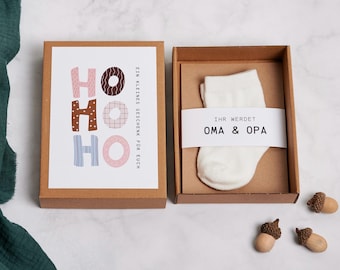 Puede incluir: Una caja de regalo con un calcetín blanco y una tarjeta con las palabras "HO HO HO" en letras rosas, marrones y azules. La caja también contiene el texto "EIN KLEINES GESCHENK FÜR EUCH" y "IHR WERDET OMA & OPA". Dos pequeñas bellotas de madera están junto a la caja.