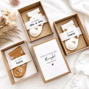 May include: Three brown cardboard gift boxes containing socks with bear patterns. Two boxes hold white socks with text bands reading IHR WERDET OMA & OPA and DU WIRST TANTE. One box holds tan socks with a band reading DU WIRST PAPA. A card reads EINE KLITZEKLEINE Kleinigkeit F&Uuml;R EUCH.