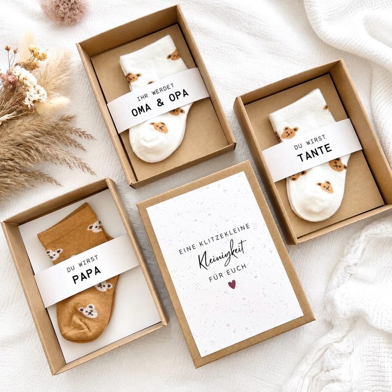 May include: Three brown cardboard gift boxes containing socks with bear patterns. Two boxes hold white socks with text bands reading IHR WERDET OMA & OPA and DU WIRST TANTE. One box holds tan socks with a band reading DU WIRST PAPA. A card reads EINE KLITZEKLEINE Kleinigkeit F&Uuml;R EUCH.