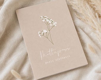 May include: A rectangular beige booklet featuring a white illustration of a sprig of baby's breath flowers. The text "Mutterpass" is printed in a white script font, followed by the name "MARIE SCHÖNBECK" in a clean white sans-serif font.