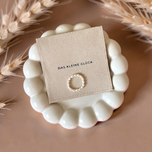Könnte beinhalten: Ein zarter Ring aus kleinen weißen Perlen, zentriert auf einer quadratischen beigen Karte. Die Karte trägt den schwarzen Aufdruck DAS KLEINE GLÜCK. Ring und Karte liegen auf einer weißen, kreisförmigen Keramikschale mit gewelltem Rand.