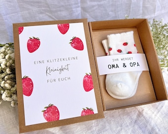 Könnte beinhalten: Eine braune Pappschachtel mit einem Paar weißer Babysocken mit kleinem Erdbeermuster und einer weißen Papierbanderole mit dem Text IHR WERDET OMA & OPA. Eine passende Karte zeigt Erdbeeren und den Text EINE KLITZEKLEINE Kleinigkeit FÜR EUCH.